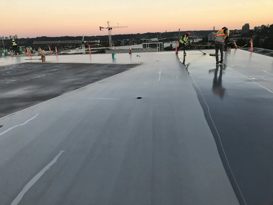 The crew spreading the grey Tremco Vulkem 350NF/950NF/951NF coat across the Royal Columbian Hospital helipad at dusk, above the Fraser River